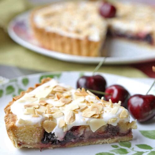 slice of cherry almond frangipane tart with cherries and tart in background.