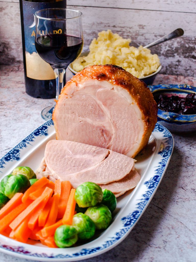 Large carved ham with carrots, brussel sprouts. Glass of wine behind the ham.