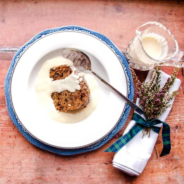 Plate with fruit pudding cake and custard. Jug of custard and napkin with heather tied to it with tartin ribbon