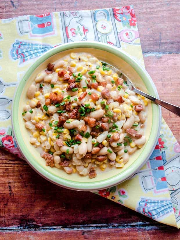 Grilled Corn with beans and bacon in a cream sauce, served in a bowl with aspoon placed on a tea towel