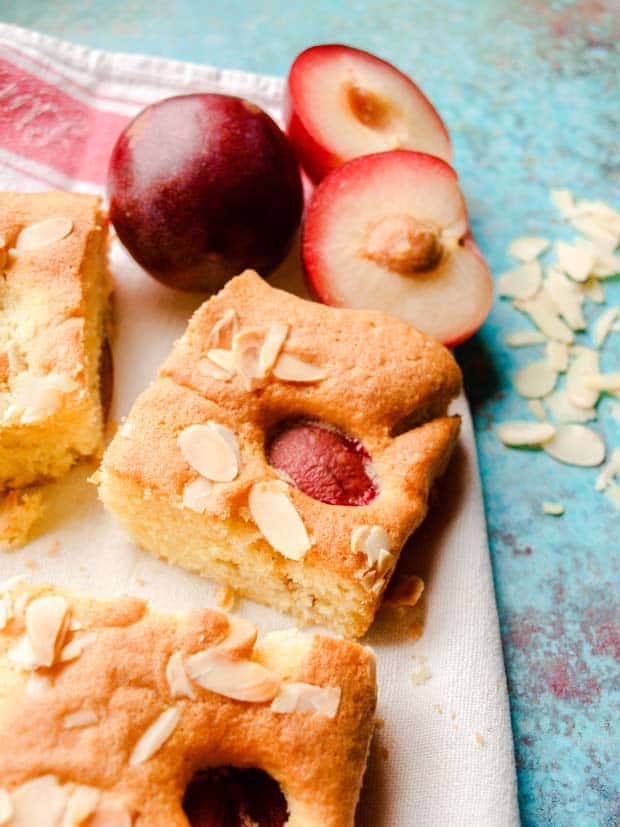 Square of Plum and Almond Traybake Cake on a tea towel with fresh plums and flaked almonds on the side.