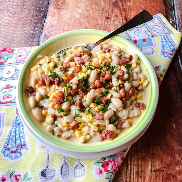 Bowl of corn, bacon and beans on a cloth with a spoon. 