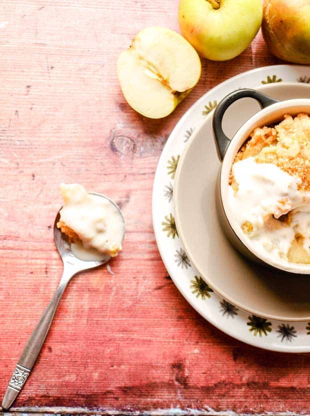 Bowl of crumble dessert with custard, spoon on wooden surface with dessert and apples.